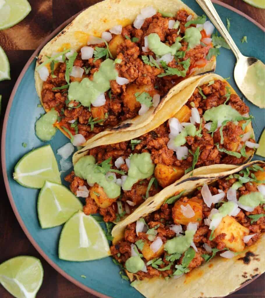 three ground pork al pastor tacos on a plate with salsa verde and lime wedges