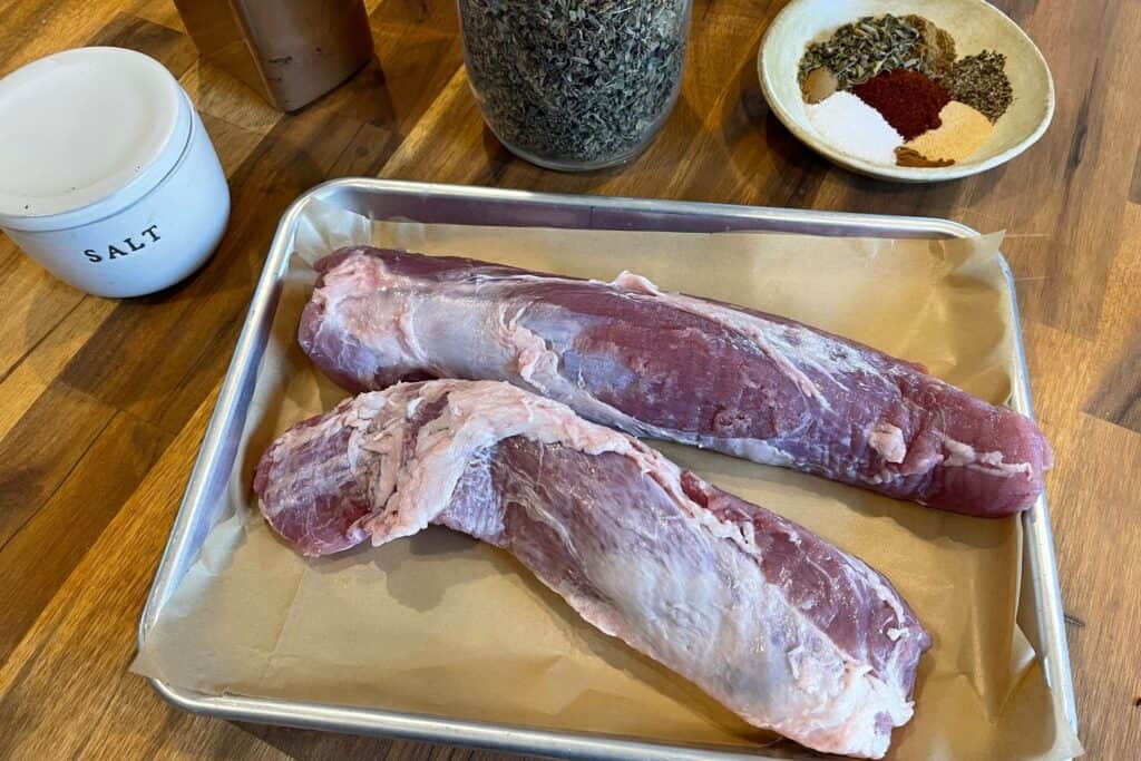 two pork tenderloins before trimming fat away next to a bowl of mixed spices