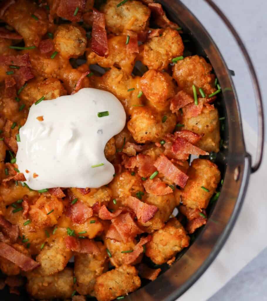 tater tots in the air fryer basket with melted cheese, bacon, sour cream, and chopped chives
