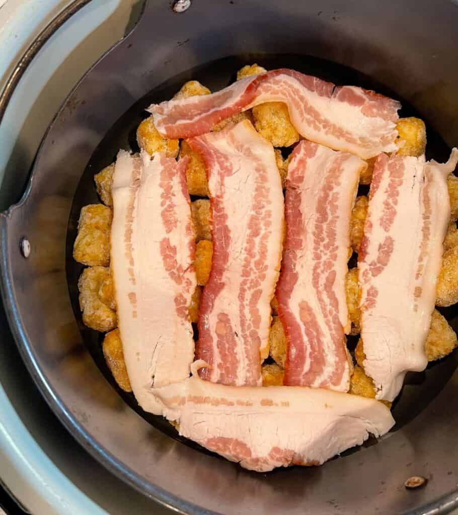 3 strips of bacon cut in half on top of frozen tater tots in an air fryer basket