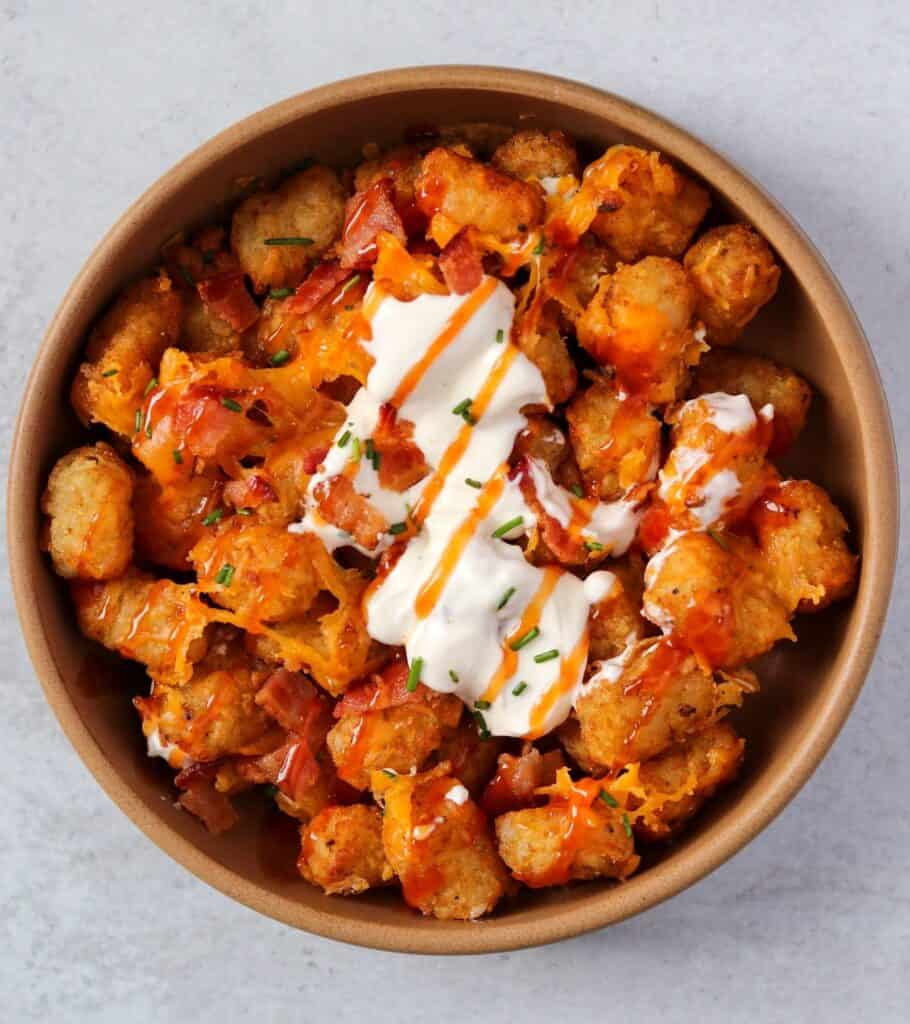 air fried tater tots in a brown bowl topped with melted cheddar, sour cream, bacon bits, and a hot sauce drizzle