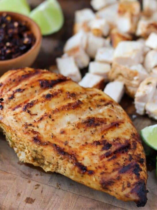 grilled chicken breast on a cutting board beside diced chicken, salsa macha, and lime wedges