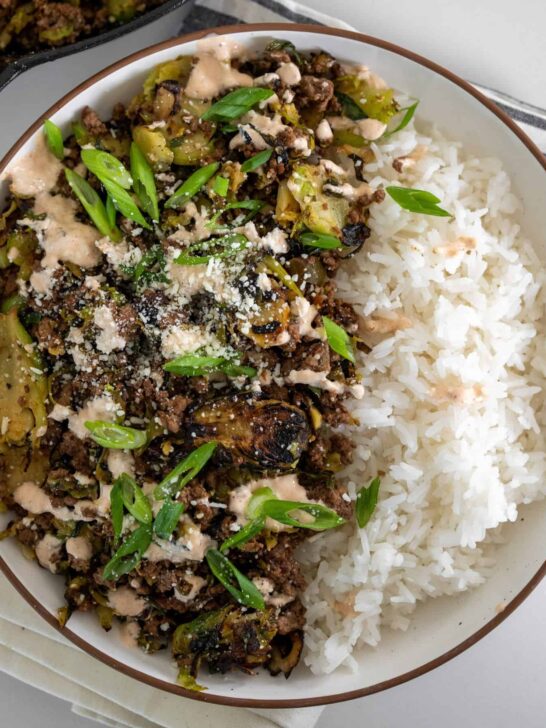 ground beef and brussels sprouts in a bowl with white rice garnished with scallions, cotija cheese, and spicy crema