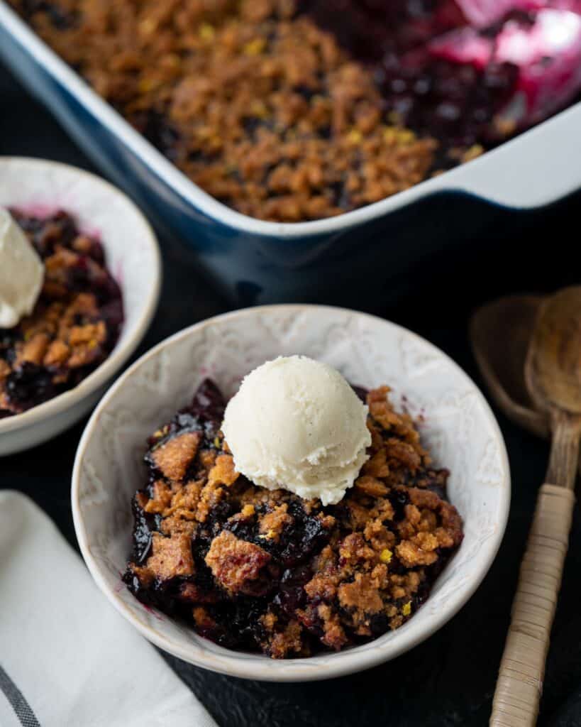 two bowls of blueberry crumble topped with vanilla bean ice cream in front of a baking dish