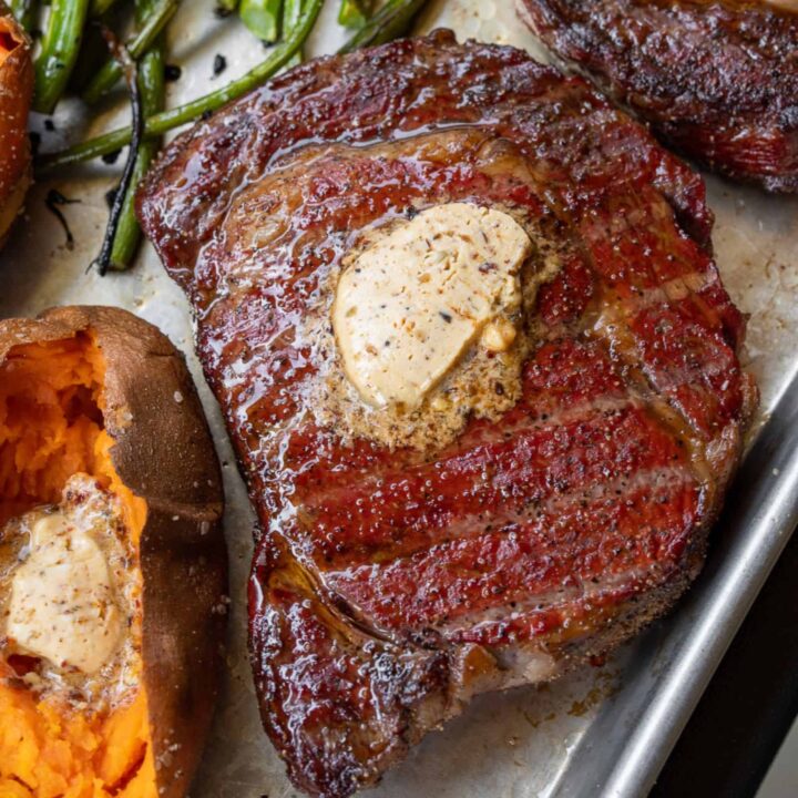 reverse seared ribeye on a sheet pan topped with compound butter next to a baked sweet potato and grilled broccolini