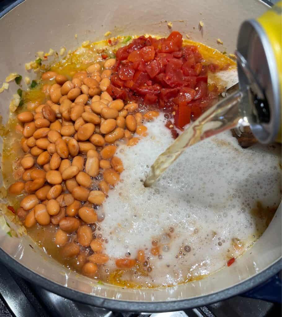 pouring beer into a Dutch oven with pinto beans, diced tomatoes, and bacon with veggies