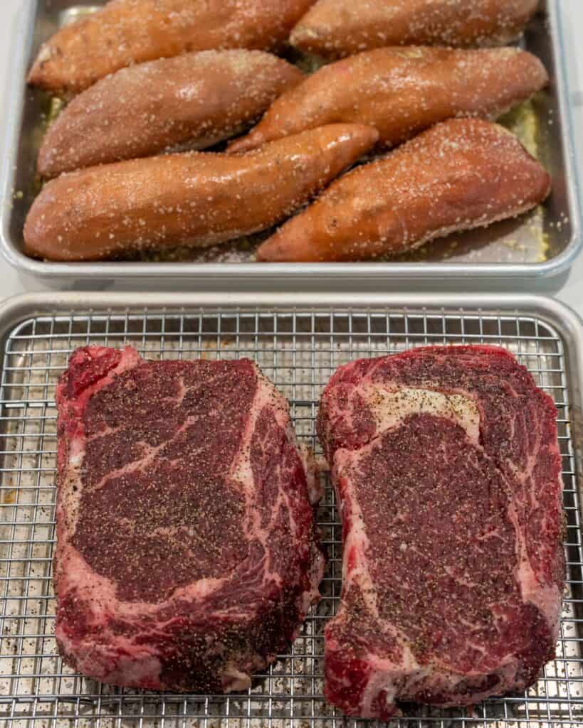 olive oil rubbed sweet potatoes on a sheet pan behind two ribeye steaks on a sheet pan with a wire rack seasoned with salt and pepper