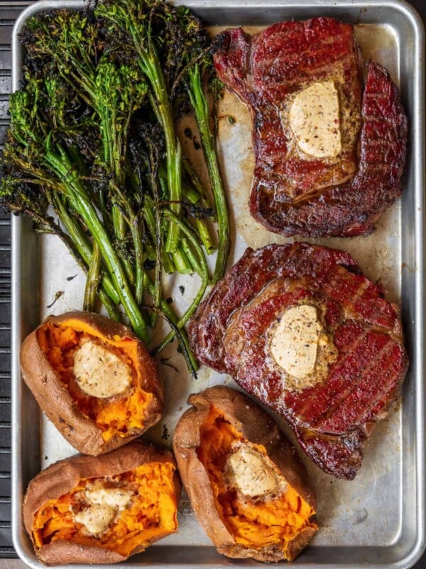 two reverse seared ribeyes on a Traeger grill with baked sweet potatoes, compound butter, and grilled broccolini