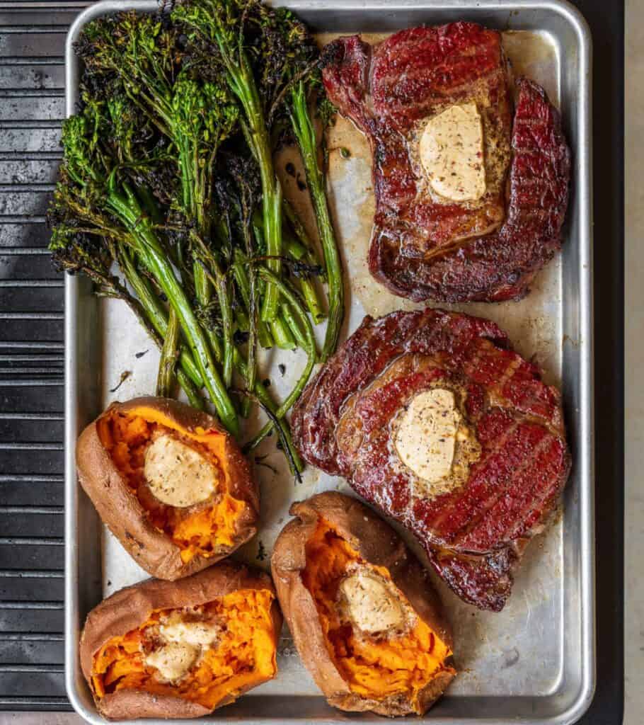 two reverse seared ribeyes on a Traeger grill with baked sweet potatoes, compound butter, and grilled broccolini
