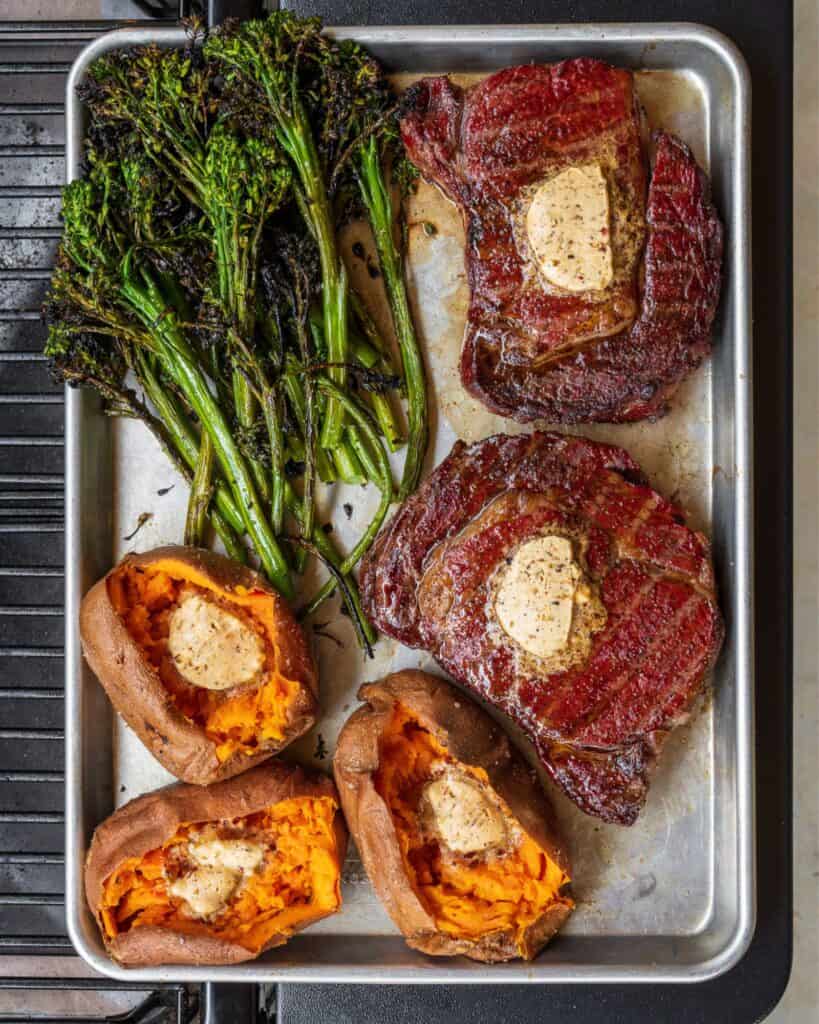 two Traeger reverse seared ribeyes on a sheet pan with baked sweet potatoes and grilled broccolini