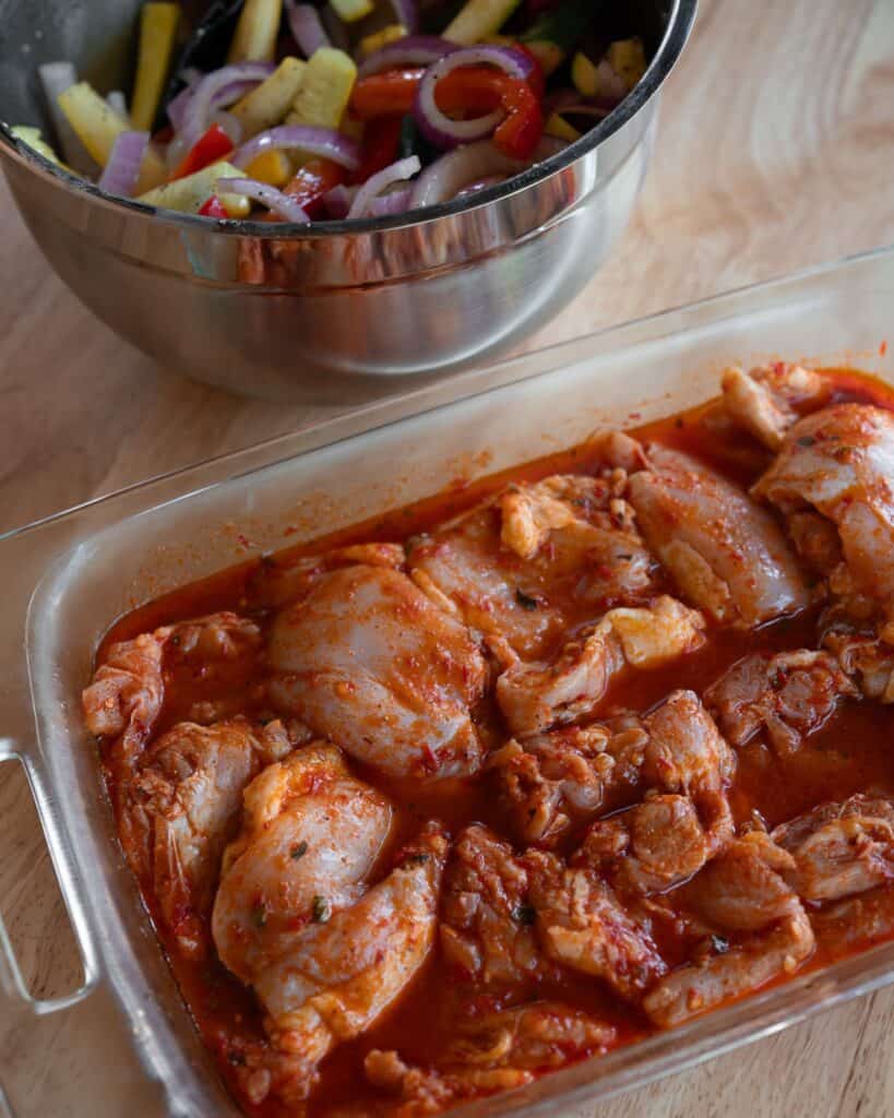 honey harissa marinated chicken thighs in a glass dish next to a mixing bowl with veggies tossed in olive oil