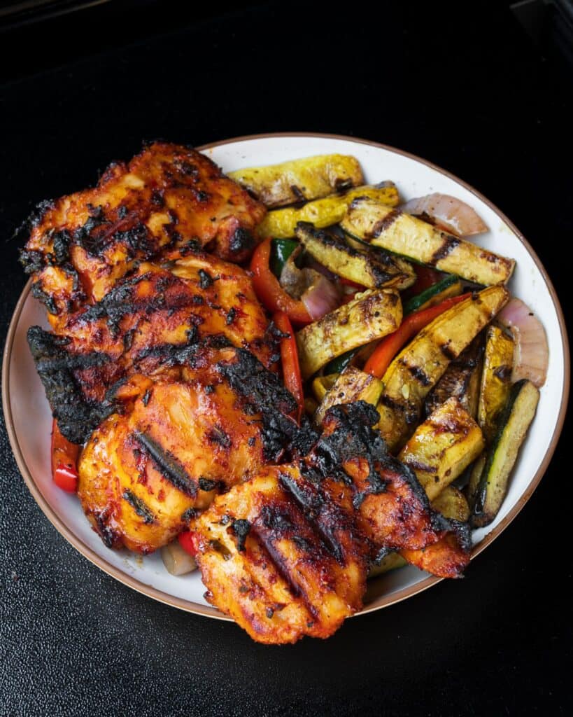 four grilled chicken thighs in a bowl with grilled vegetables