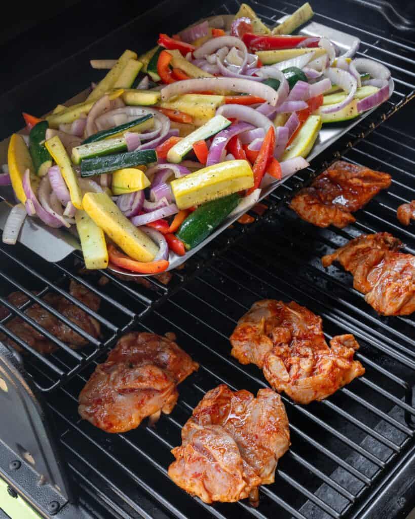 yellow squash, zucchini, peppers, and onion on a grill pan above boneless chicken thighs on the bottom rack of a Traeger grill